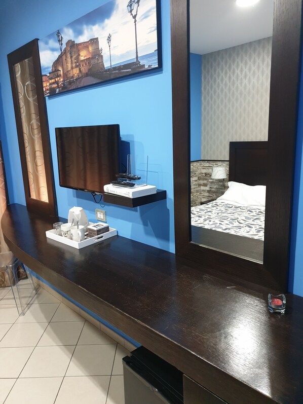 Una habitación de hotel con un escritorio de madera oscura, un televisor de pantalla plana, un estante pequeño con un control remoto y una cafetera, y una cama con una colcha estampada.