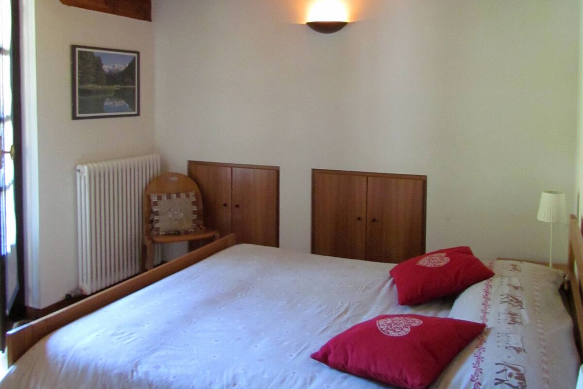 Een slaapkamer met een houten plafond, een bed met een witte dekbedovertrek en twee rode kussens, een houten hoofdbord, een houten kledingkast en een wandlamp.