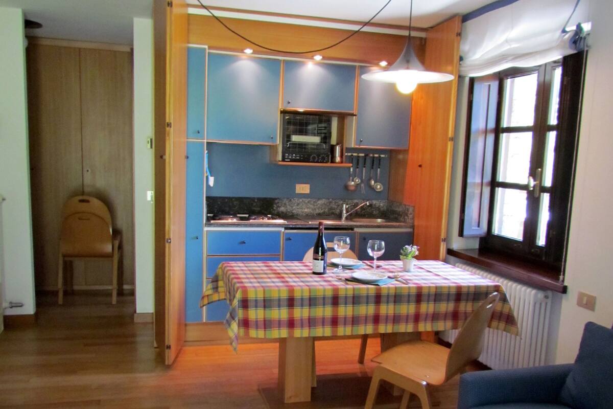 Een kleine keuken met blauwe kastjes, een eettafel met geruit tafelkleed en een blauwe bank.