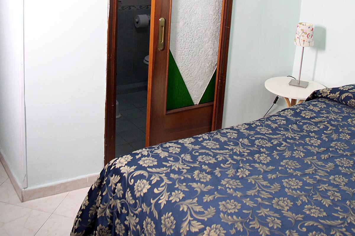 A bedroom with a wooden door, a bed with a floral bedspread, a small round table with a lamp, and a green and white geometric design on the door.