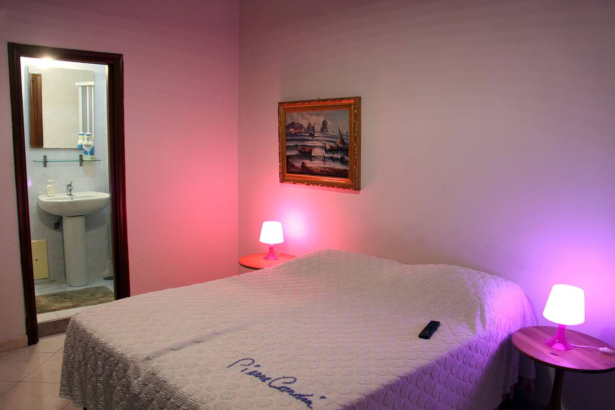 A hotel room with a bed, a bedside table with a lamp, a painting on the wall, and a bathroom visible through an open door.