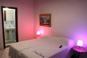 A hotel room with a bed, a bedside table with a lamp, a painting on the wall, and a bathroom visible through an open door. - B&B Cinquestelle (Naples)