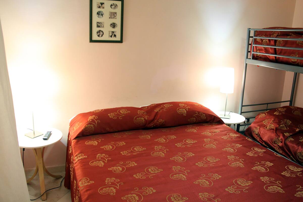 A single bed with a red patterned bedspread, a bedside table with a lamp, and a framed picture on the wall.