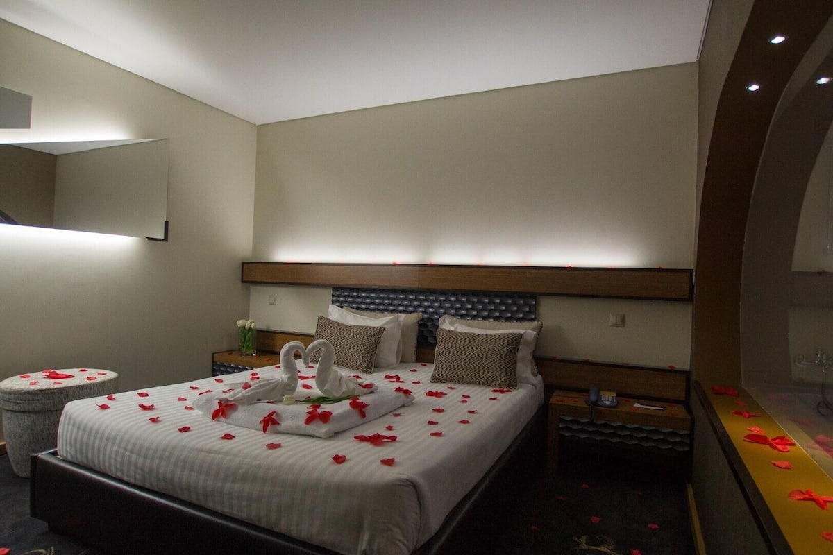 Une chambre d’hôtel avec un grand lit, une table de chevet, une commode, un miroir et une baignoire remplie de pétales de rose.