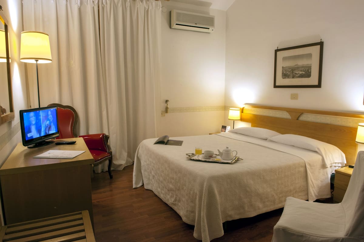 Ein Hotelzimmer mit einem Bett, einem Schreibtisch mit Computer, einem Stuhl, einer Lampe und einem Bild an der Wand.