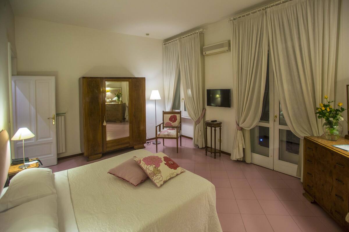 Ein Hotelzimmer mit Bett, Sessel, Schreibtisch, Spiegel, Fernseher und einem Fenster mit Vorhängen.
