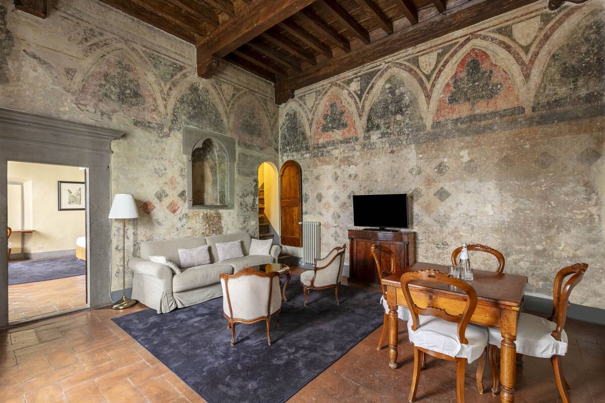 Una sala de estar con un sofá, sillas, una mesa y un televisor. El piso está cubierto de baldosas y hay una chimenea.