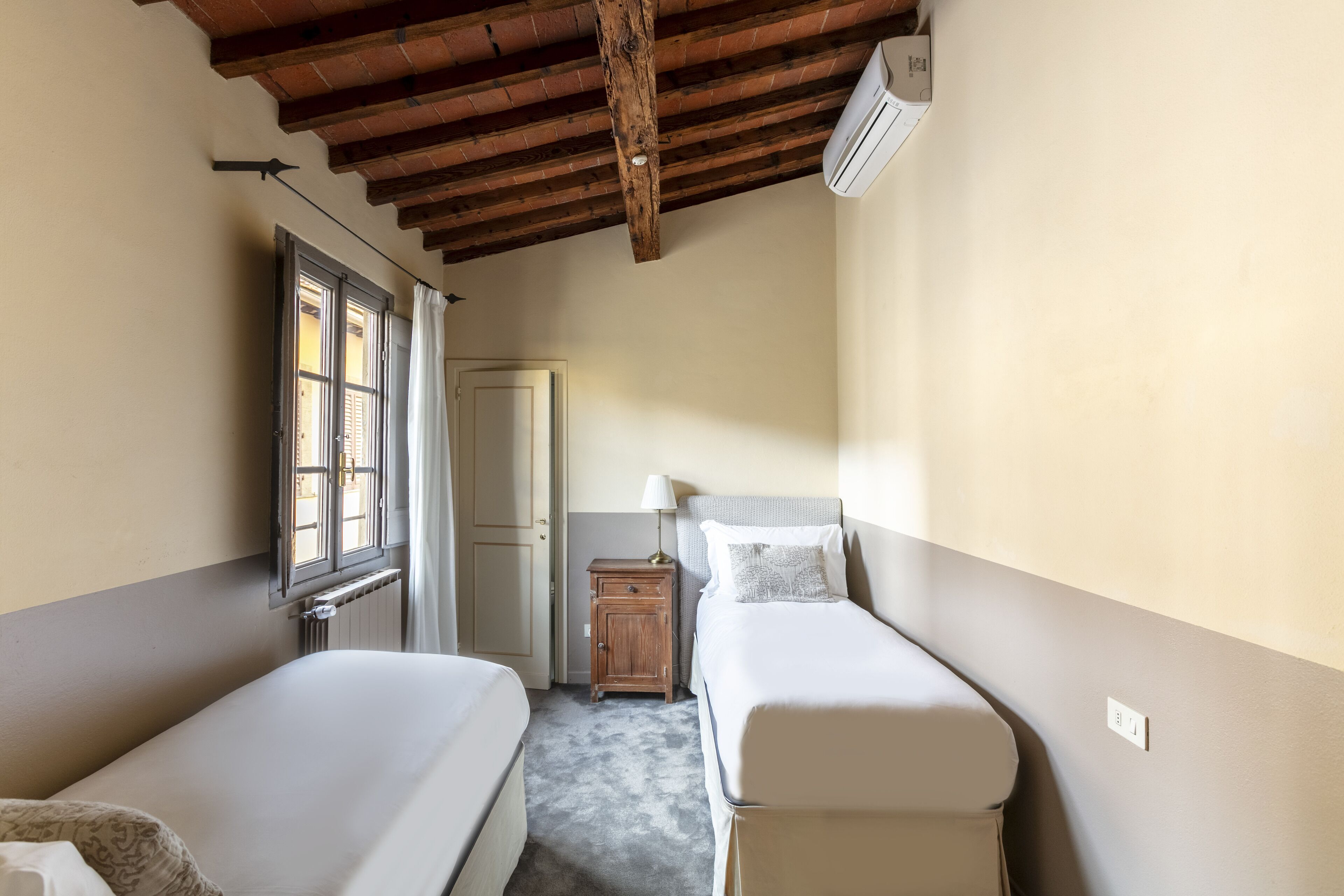 A room with two single beds, a wooden ceiling, an air conditioning unit, a small wooden cabinet, and a window with curtains.