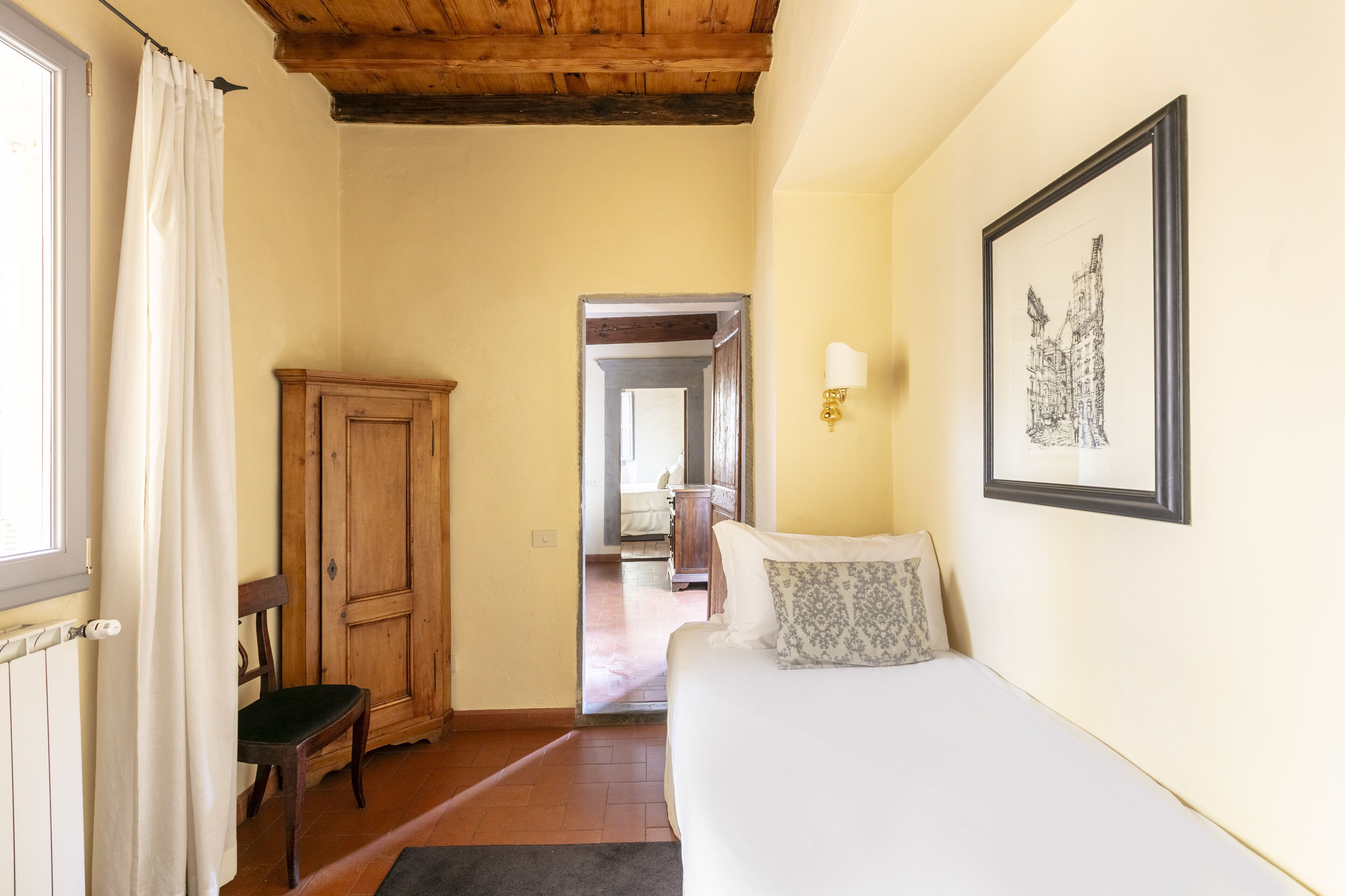 A bedroom with a bed, a wooden wardrobe, a chair, a framed picture on the wall, and a doorway leading to another room.
