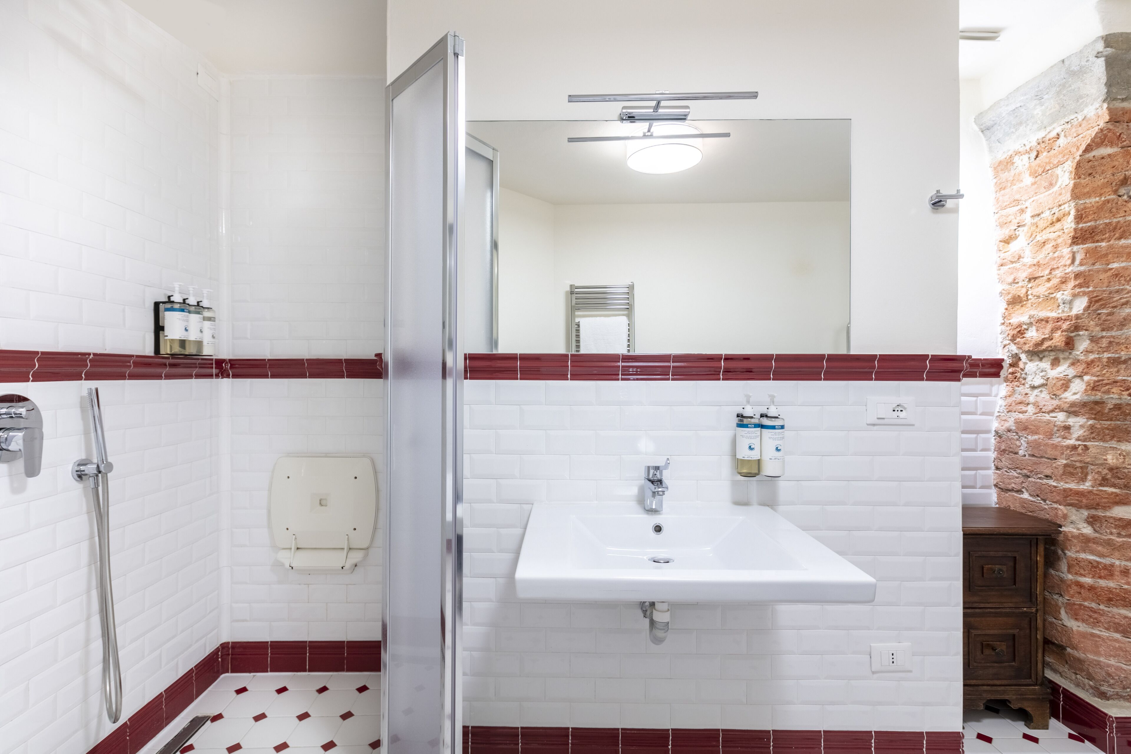 A bathroom with white tiled walls, a red-tiled floor, a white sink with soap dispensers, a shower, and a wooden cabinet.