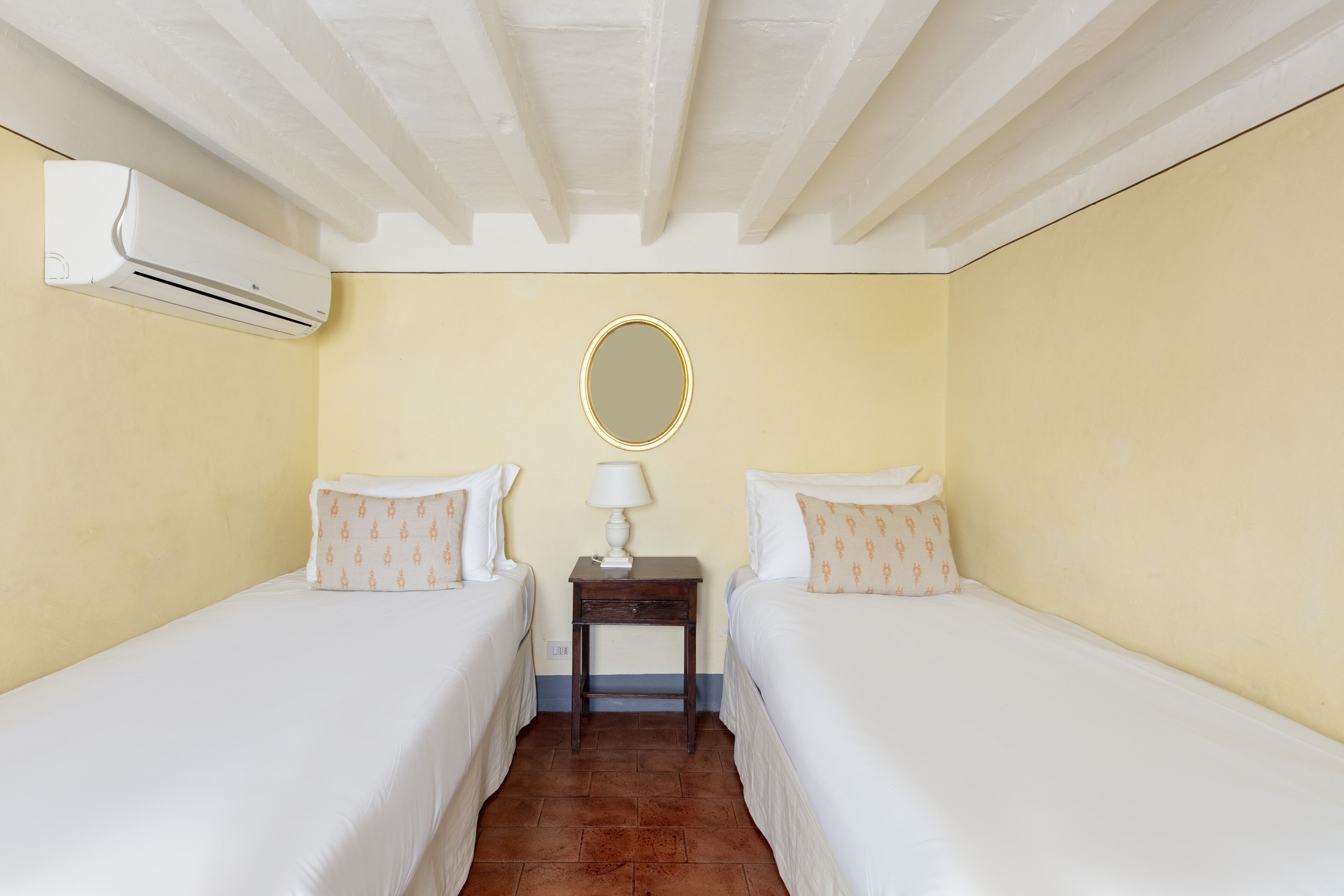 A hotel room with two single beds, a small table with a lamp, an air conditioning unit, and a round mirror on the wall.