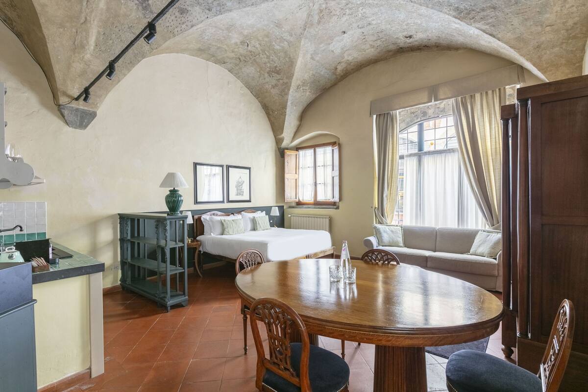 Una habitación con una mesa redonda de madera, una cama con ropa blanca, un sofá y una kitchenette.