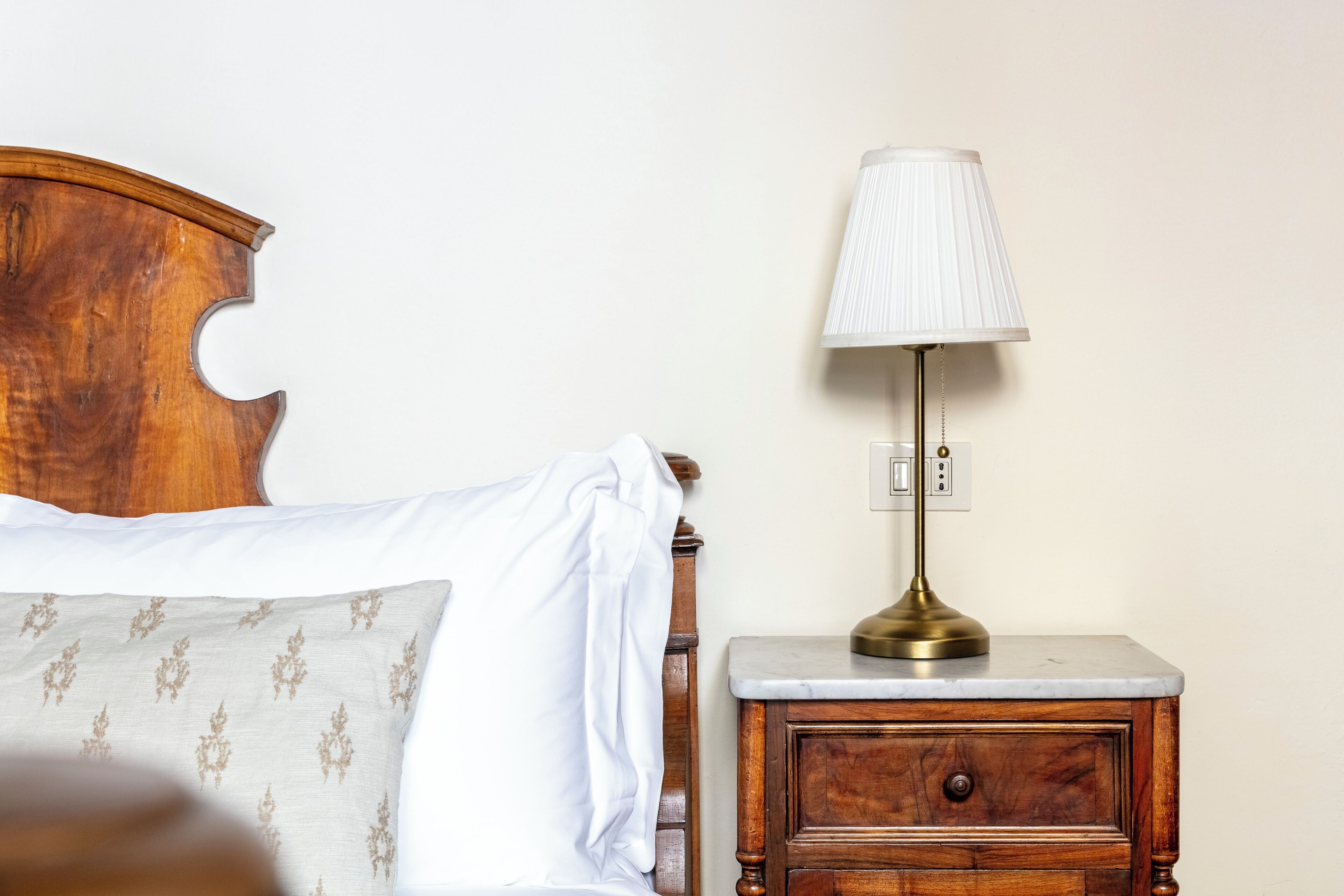 A wooden nightstand with a brass lamp, a white bedside table, and a bed with white bedding and patterned pillows.