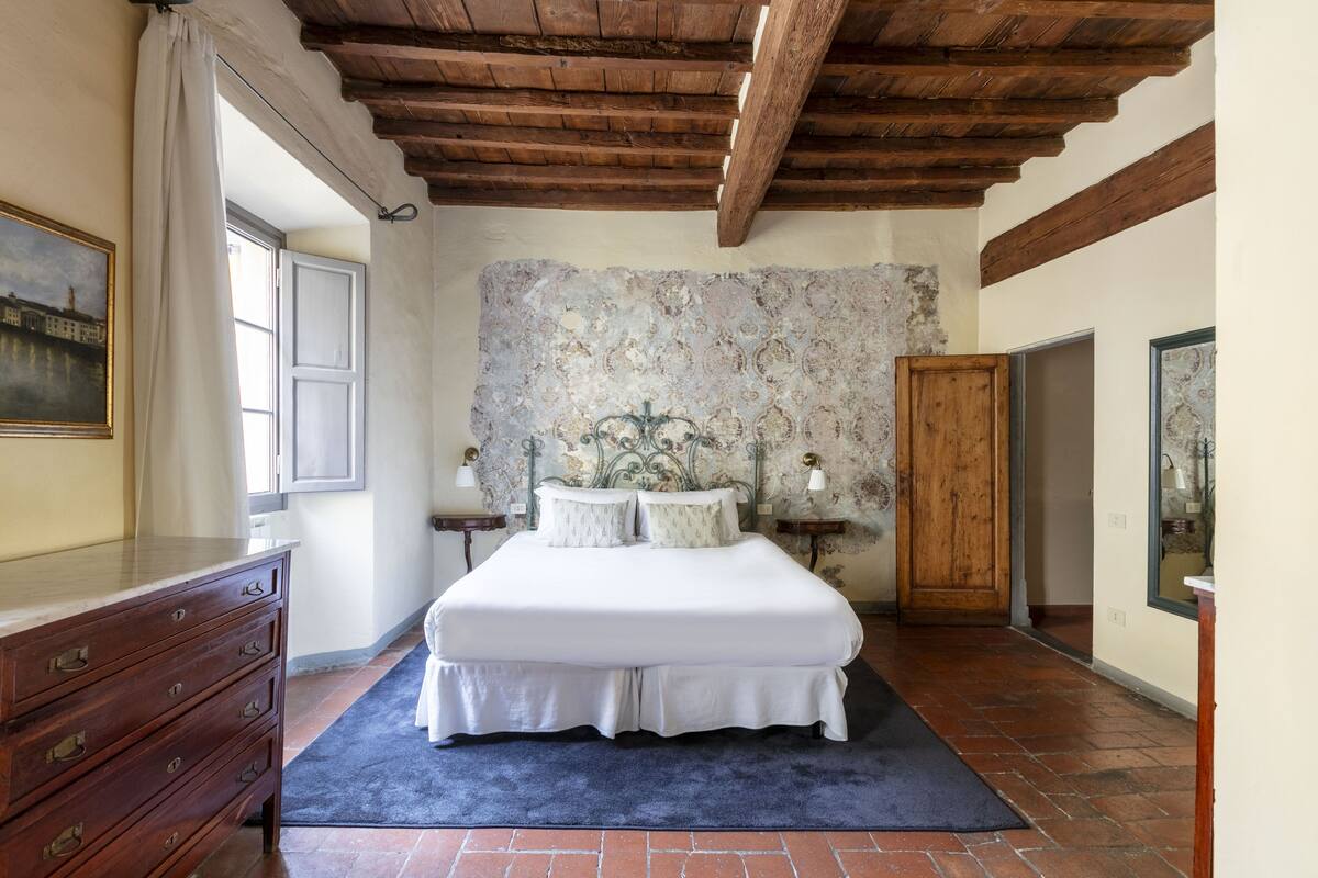 Un dormitorio con techo de madera, una cama con ropa blanca, un aparador y un ventanal grande.