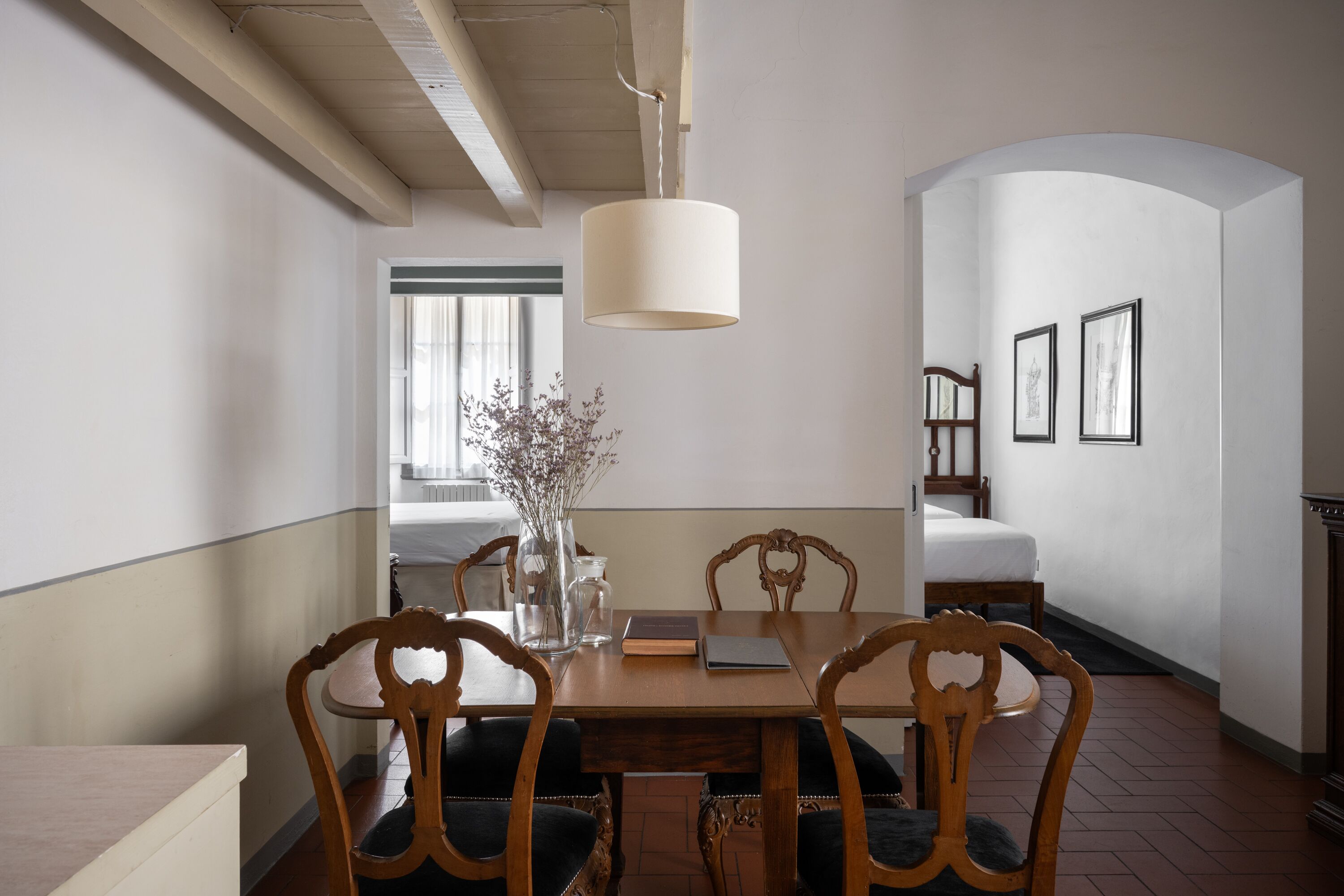 A dining area with a wooden table, four chairs, a vase with flowers, a book, and a hanging light fixture.