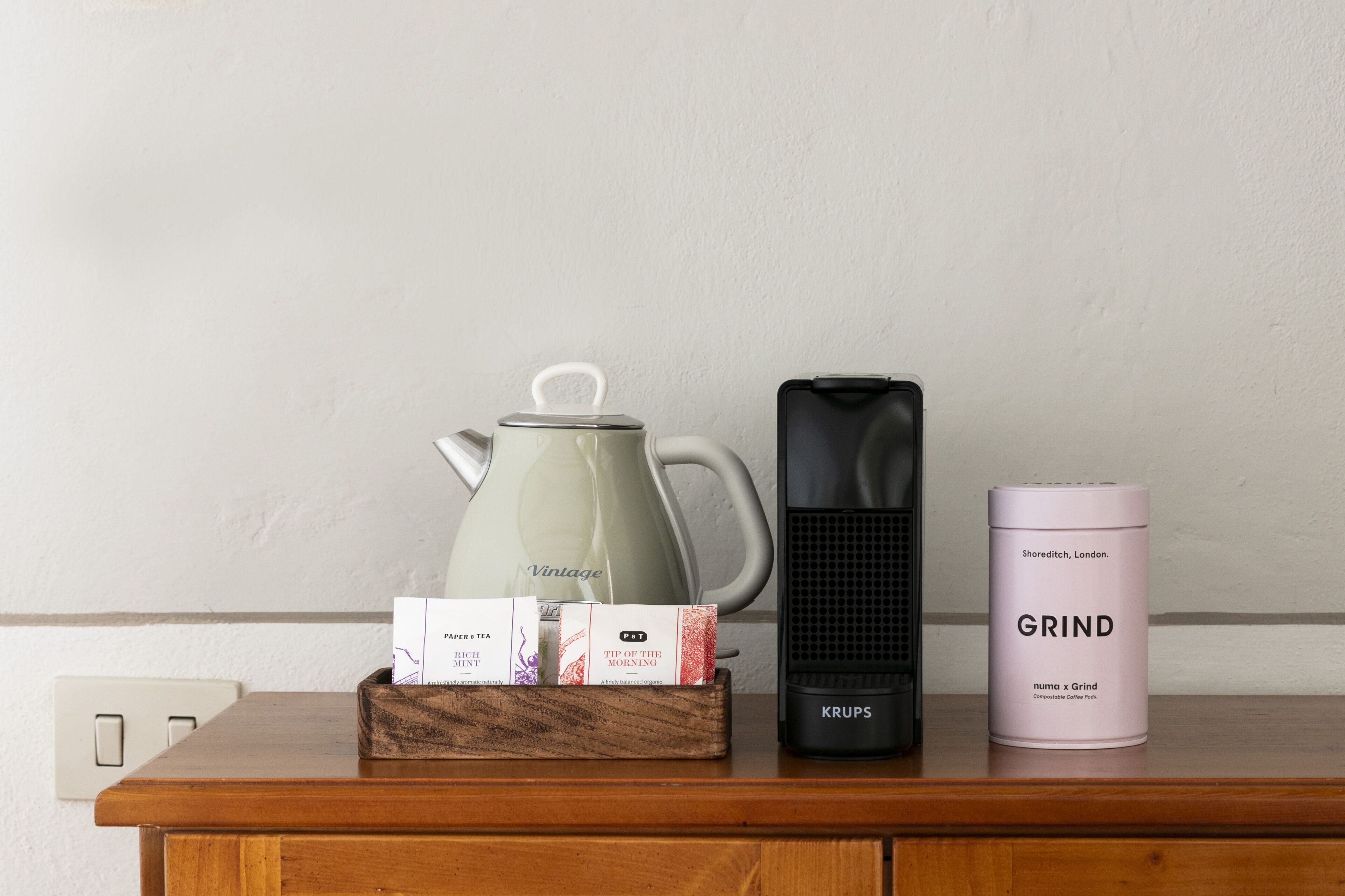 A wooden table with a kettle, a portable heater, a box of tea, and a container of coffee.