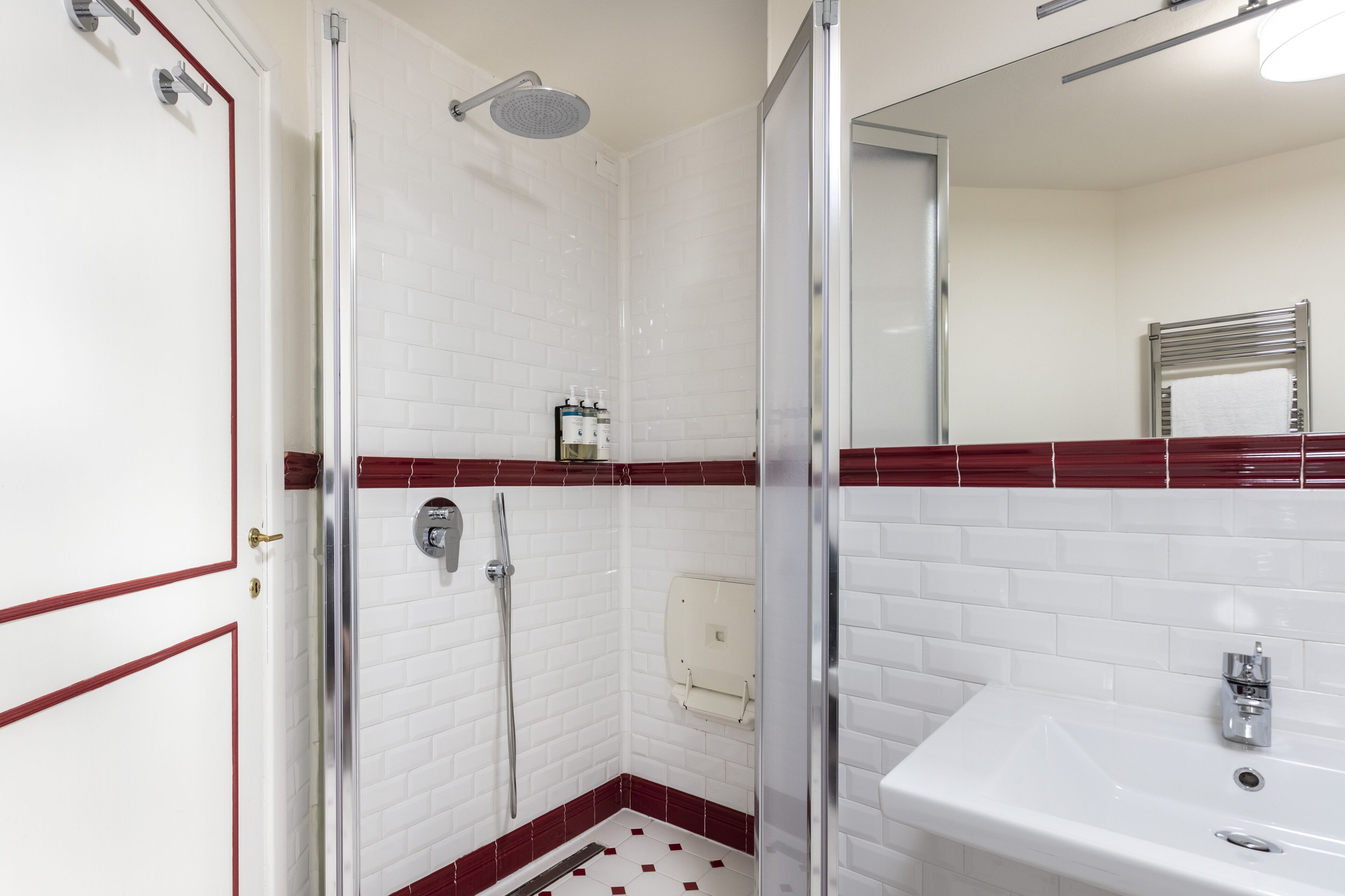 Une salle de bain avec des murs recouverts de carreaux blancs, un sol carrelé de rouge, une douche, un lavabo et un miroir.
