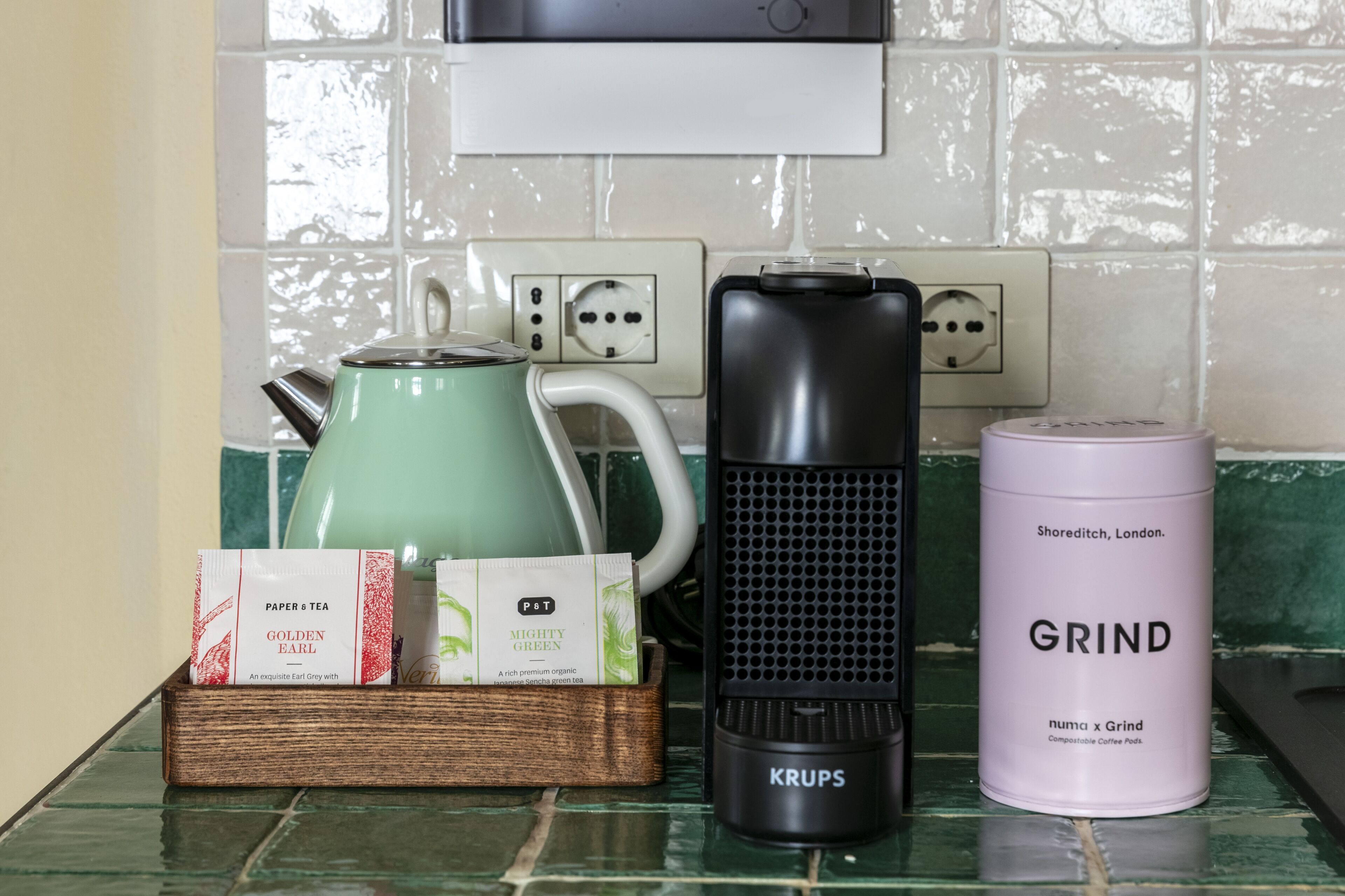 A kitchen counter with a kettle, a coffee maker, a coffee pod container, and a coffee grinder.