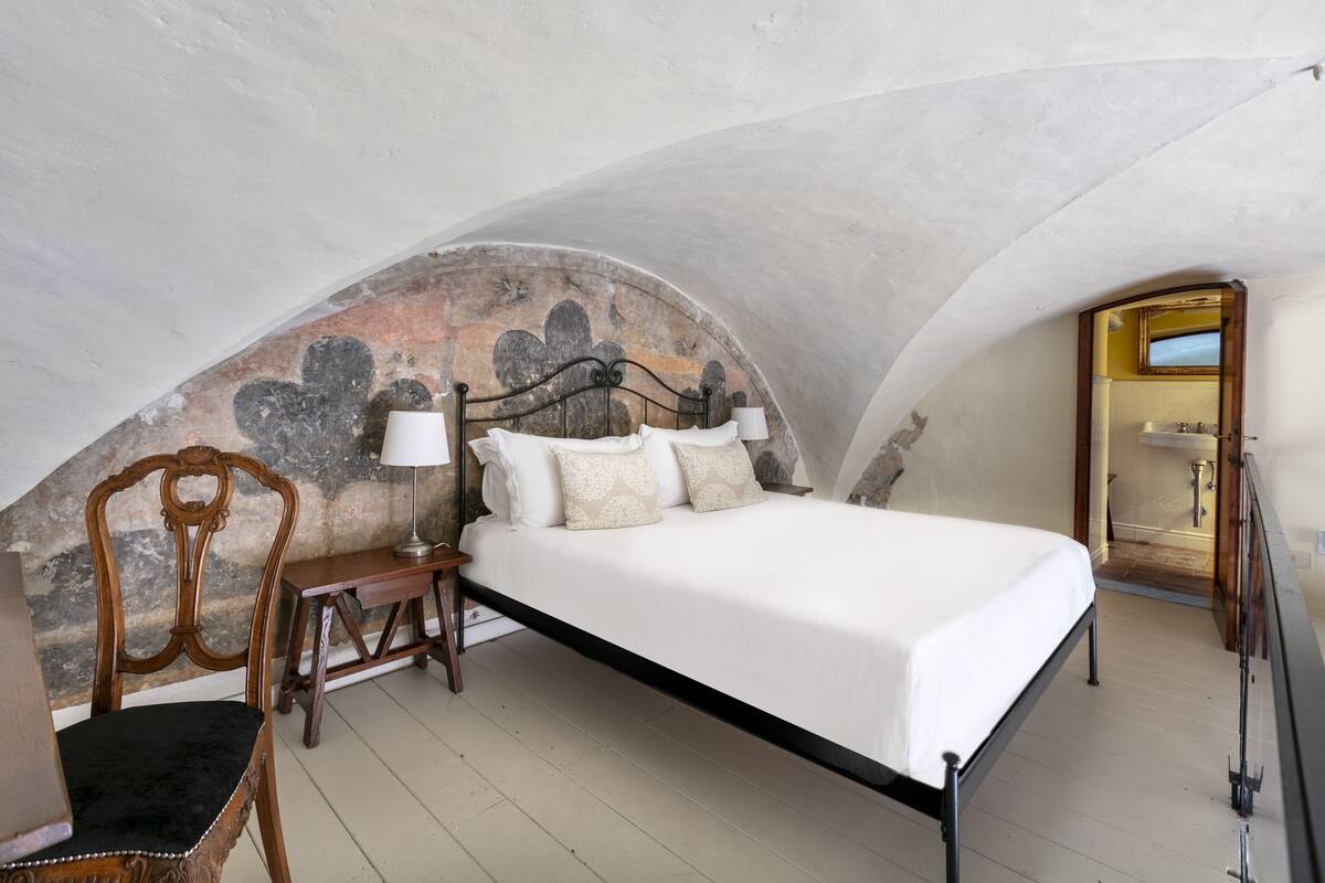 Un dormitorio con cama, una silla, una mesita de noche y un baño visible a través de un arco.