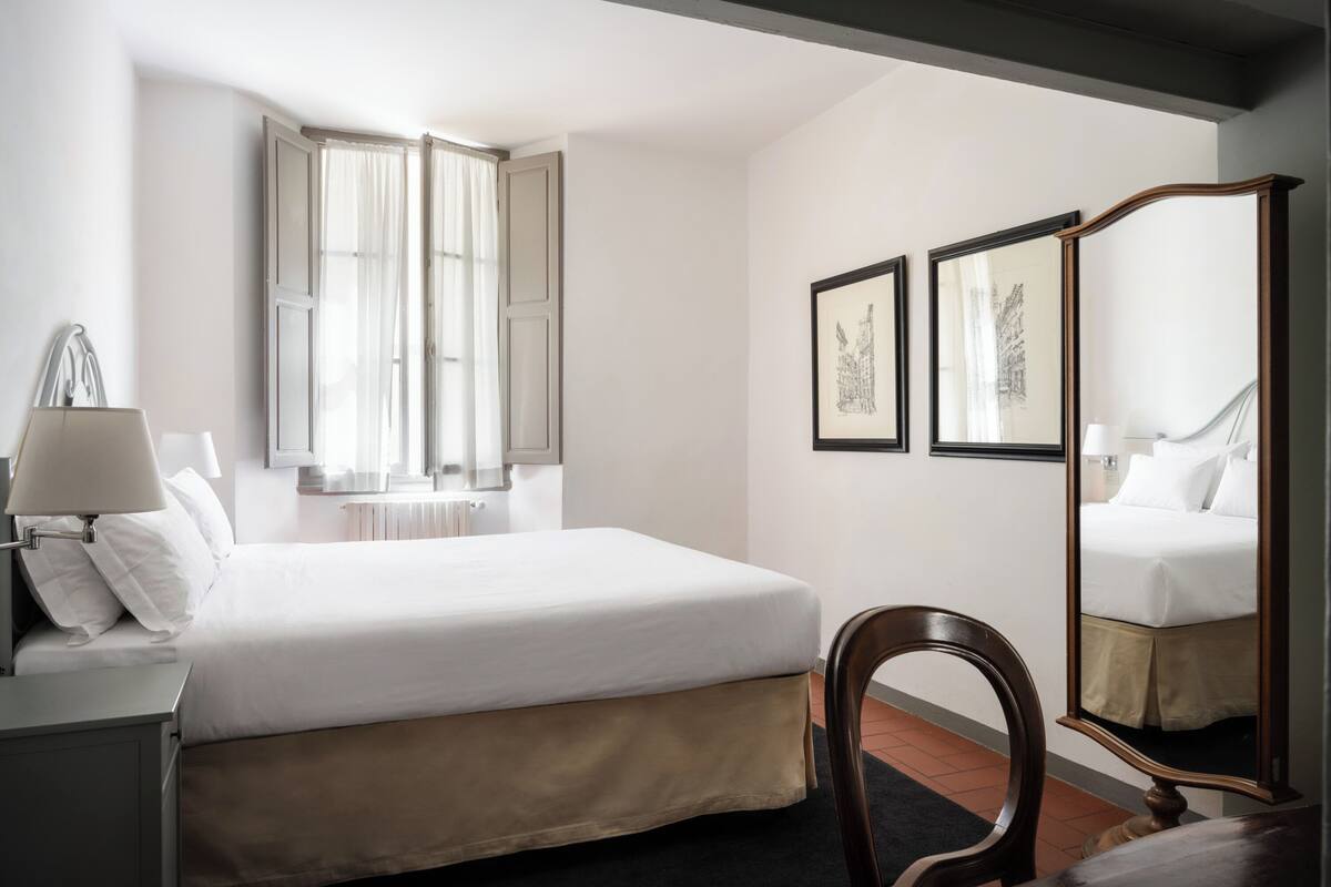 Habitación de hotel con cama, una silla, escritorio, lámpara, ventana con cortinas y cuadros en la pared.