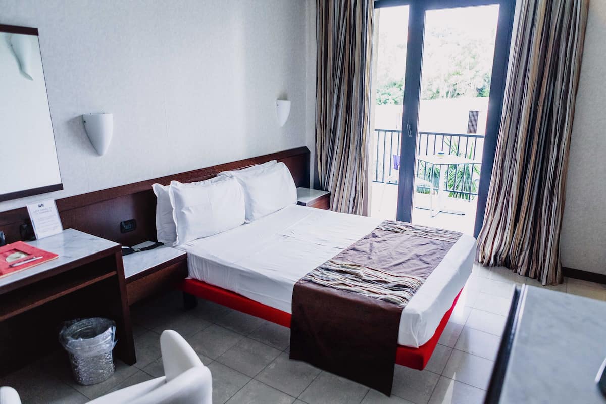 A hotel room with a bed, a desk, a chair, a trash bin, and a window with curtains.
