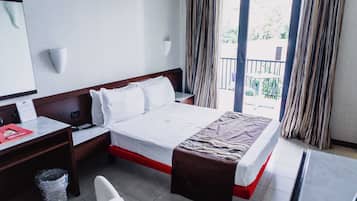 A hotel room with a bed, a desk, a chair, a trash bin, and a window with curtains.