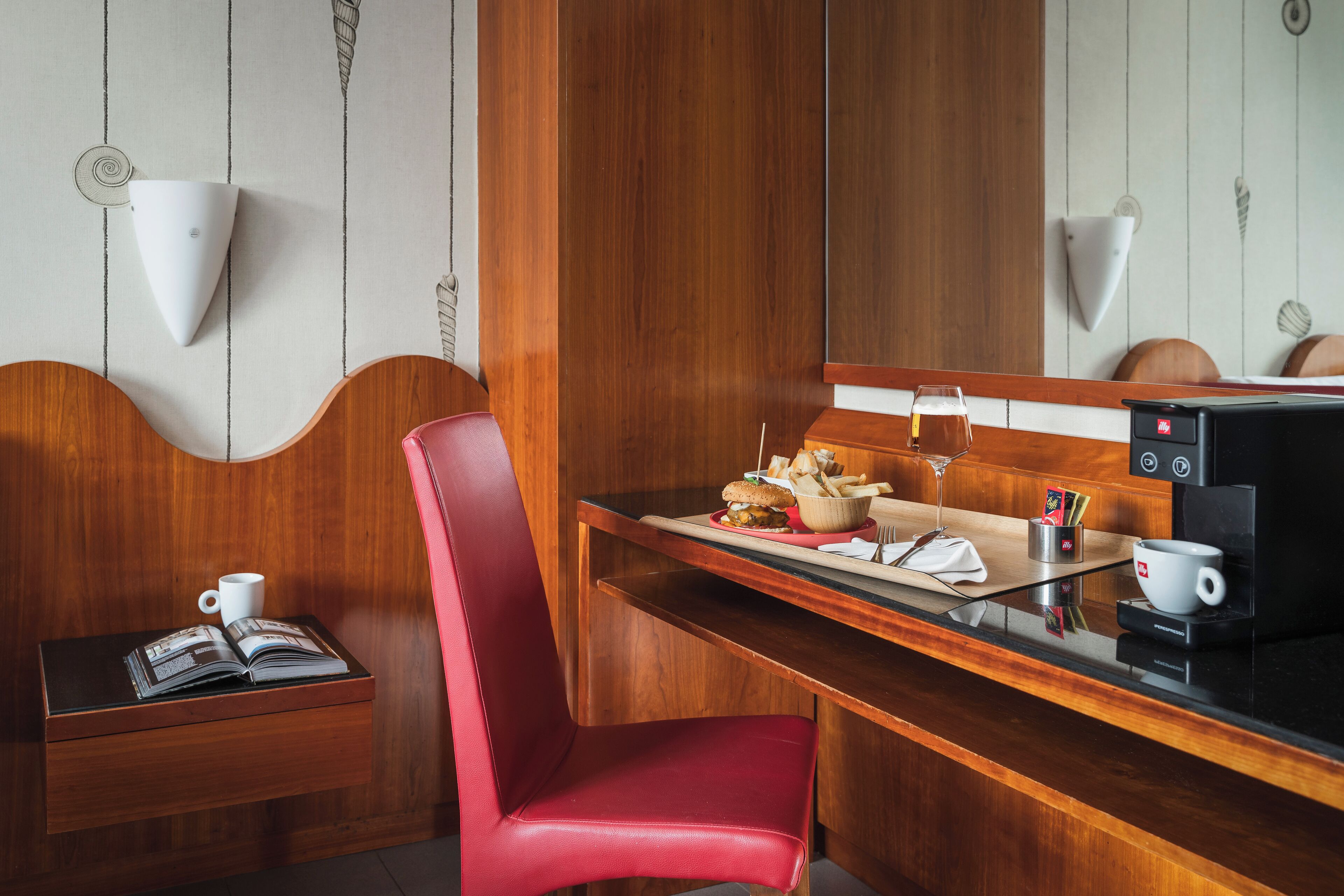 Une chambre d’hôtel avec un bureau en bois, une chaise rouge, une machine à café et un petit coin repas comprenant un panier de nourriture et un verre de bière.