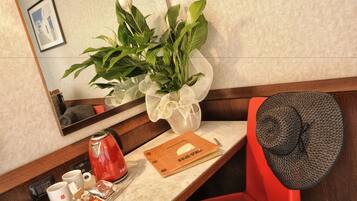 Habitación de hotel con escritorio de madera, hervidor rojo, bandeja con tazas de café y una caja de pañuelos, un cuadro en la pared y una planta en maceta.