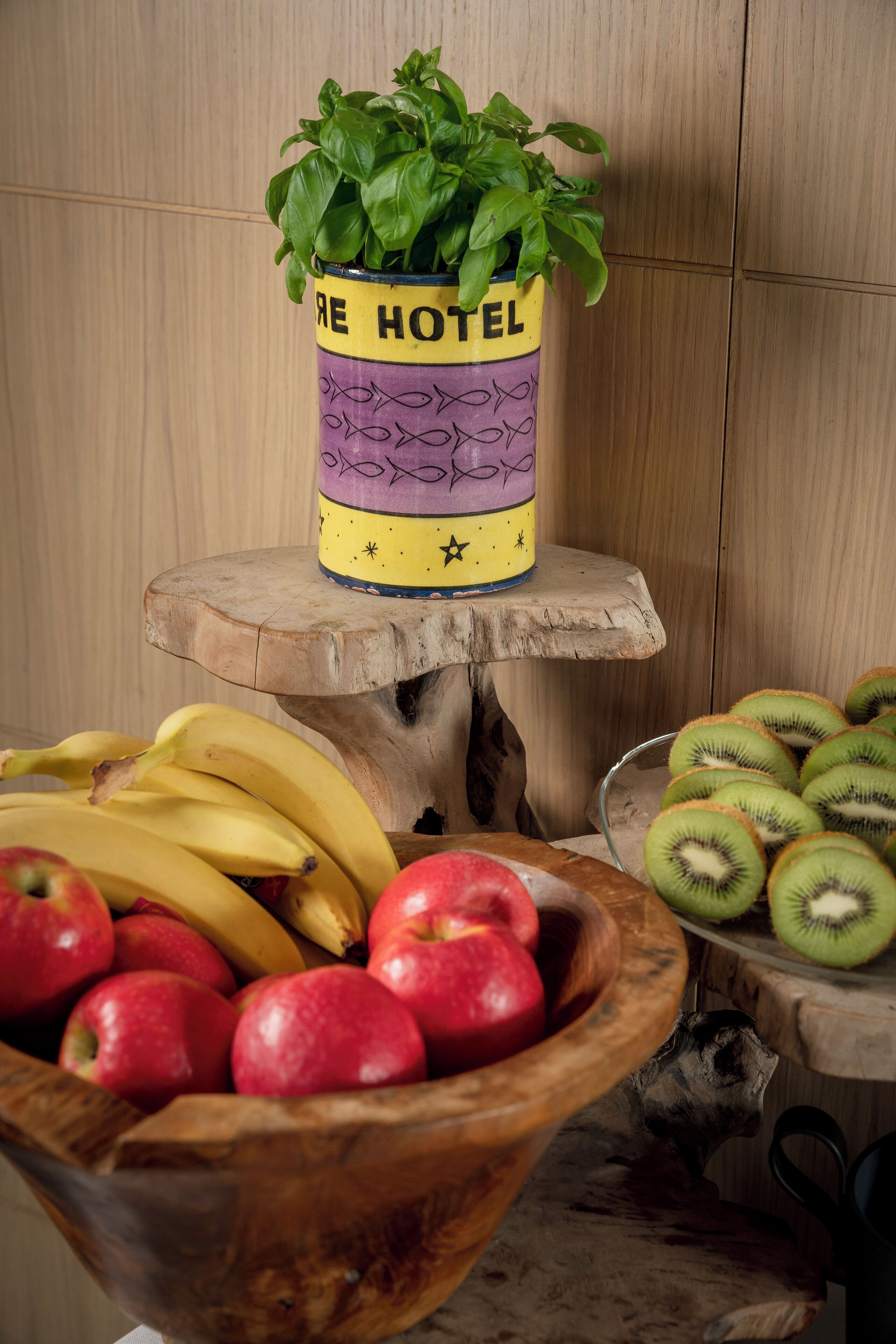 A wooden table with a bowl of apples and bananas, a bowl of kiwi, and a potted basil plant on a wooden stand.