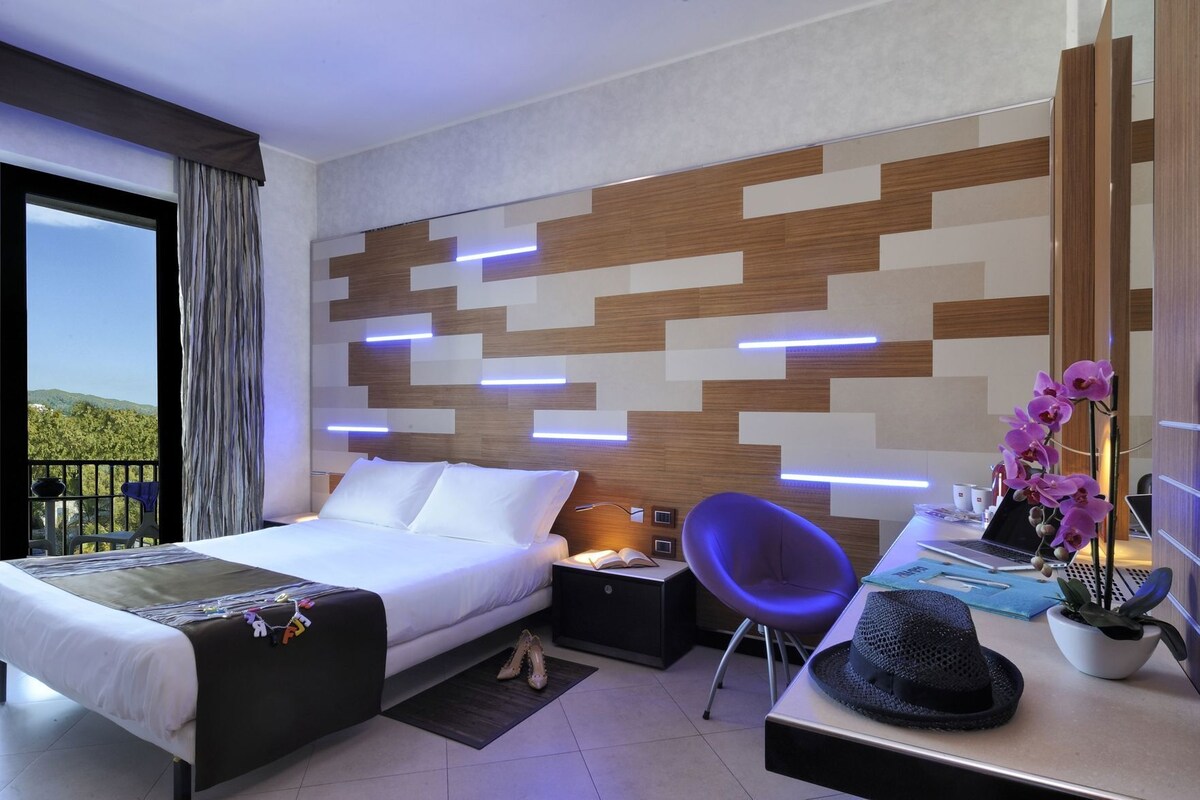 A modern hotel room with a bed, a desk with a laptop, a chair, a nightstand, and a view of the outdoors.
