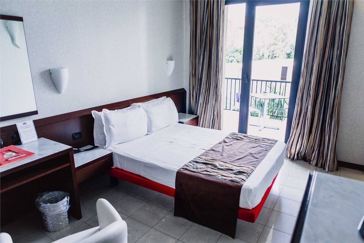 A hotel room with a bed, bedside tables, a desk, a chair, a trash bin, and a view of the outdoors through a balcony.