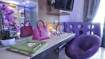 Una reception con una sedia viola, una borsa rosa, una pianta in vaso e un televisore montato a parete.