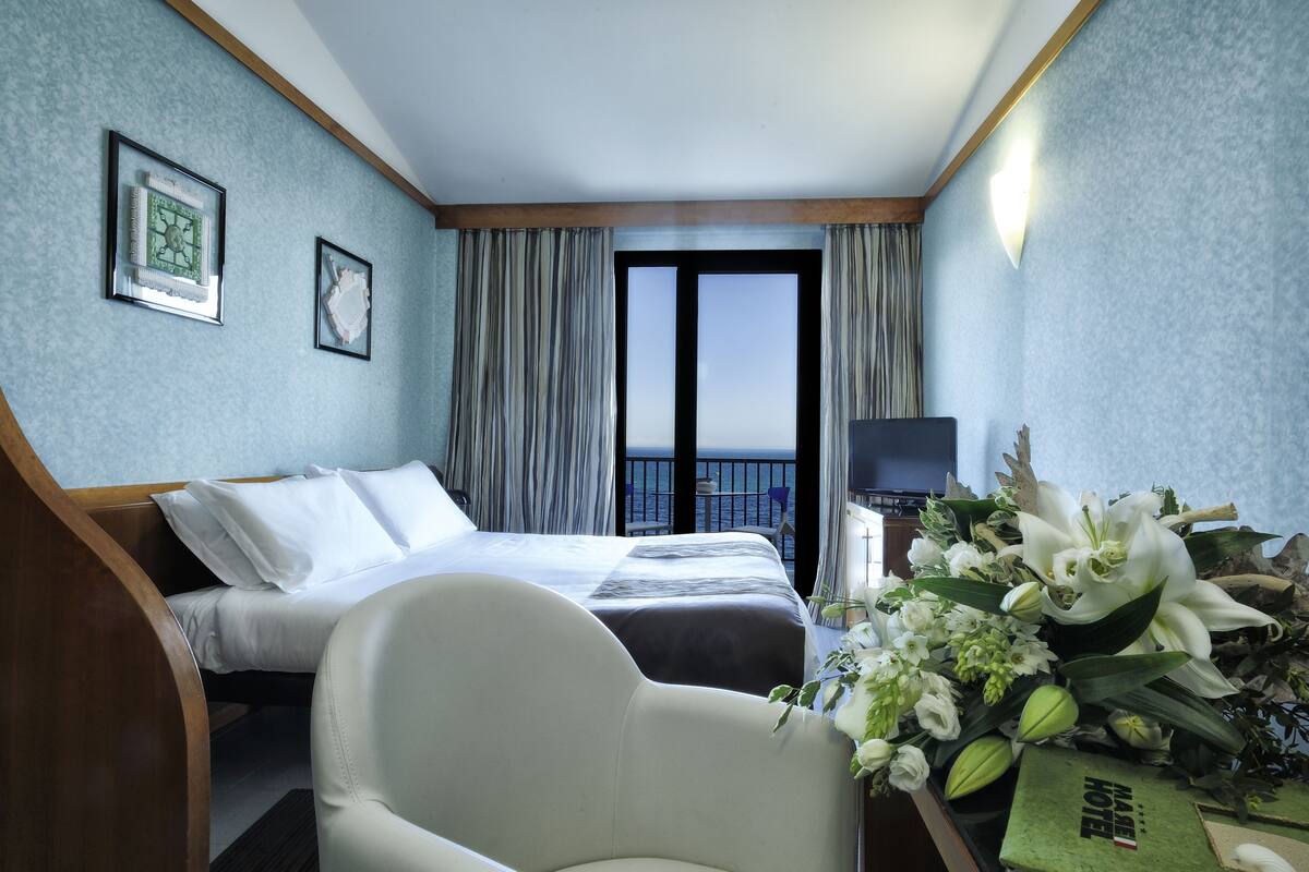 A hotel room with a bed, a chair, a desk with a lamp, a TV, and a view of the sea through a large window.