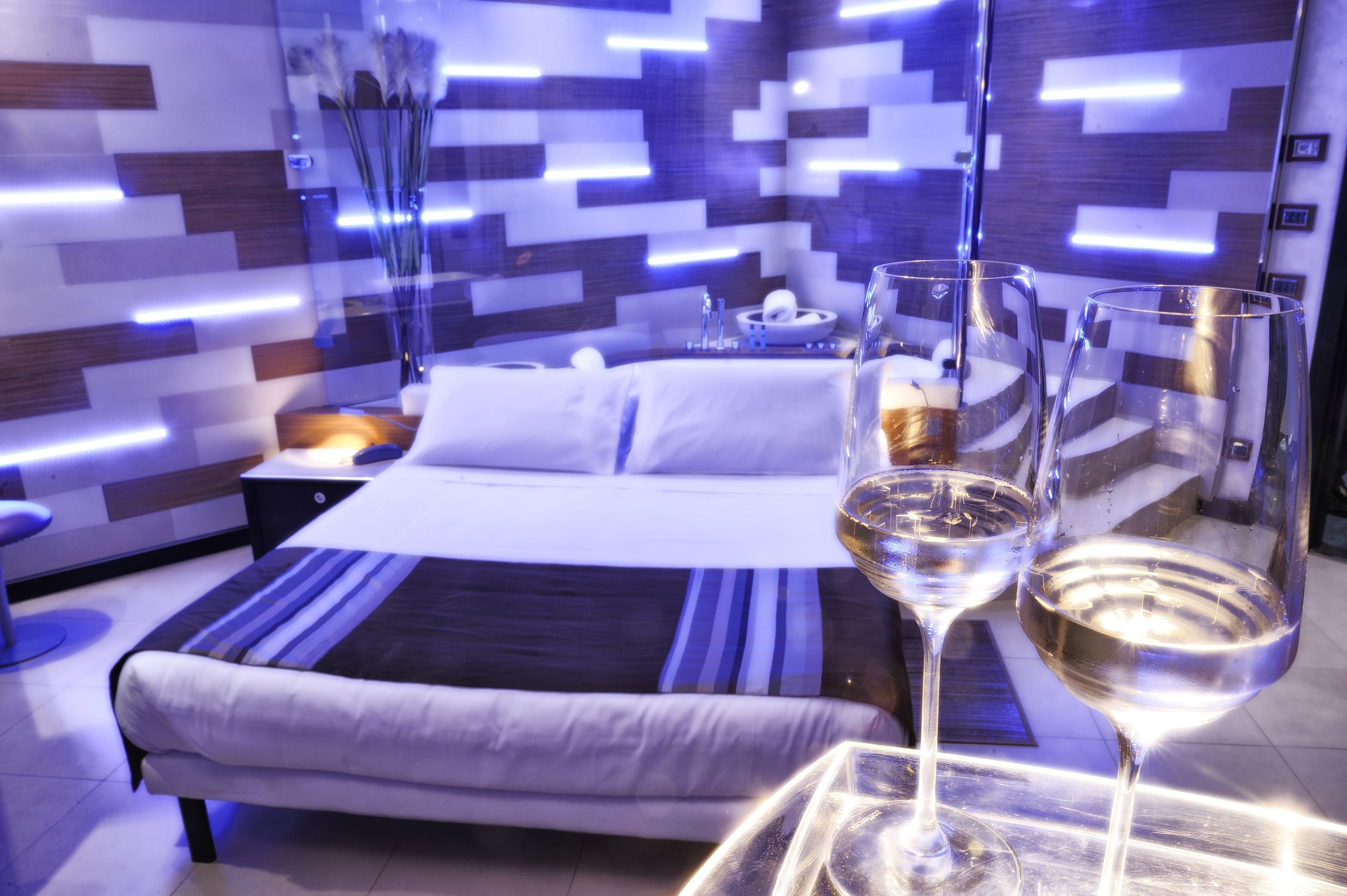 Una camera d'hotel moderna con un letto rifatto, un comodino con una lampada e due bicchieri da vino su un tavolino.