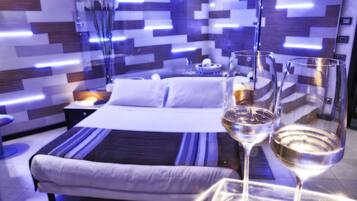 Una camera d'hotel moderna con un letto rifatto, un comodino con una lampada e due bicchieri da vino su un tavolino.