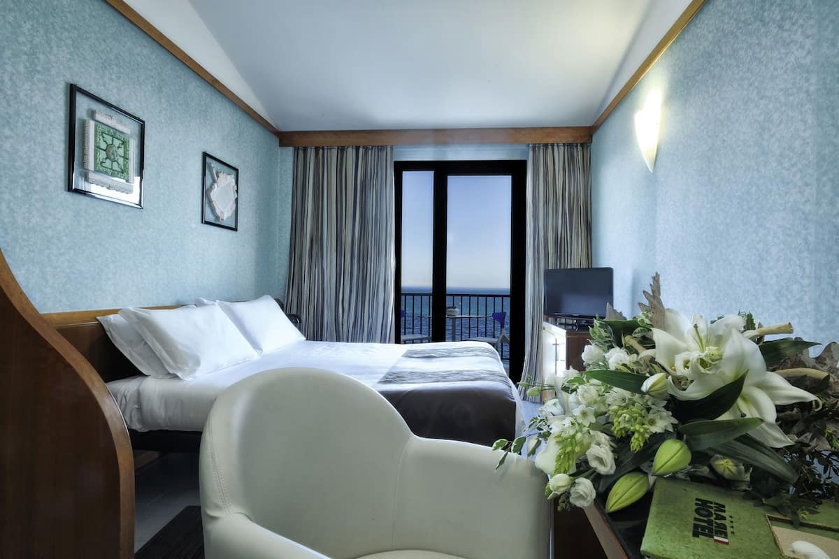 A hotel room with a bed, a desk with a TV, a chair, and a view of the sea.