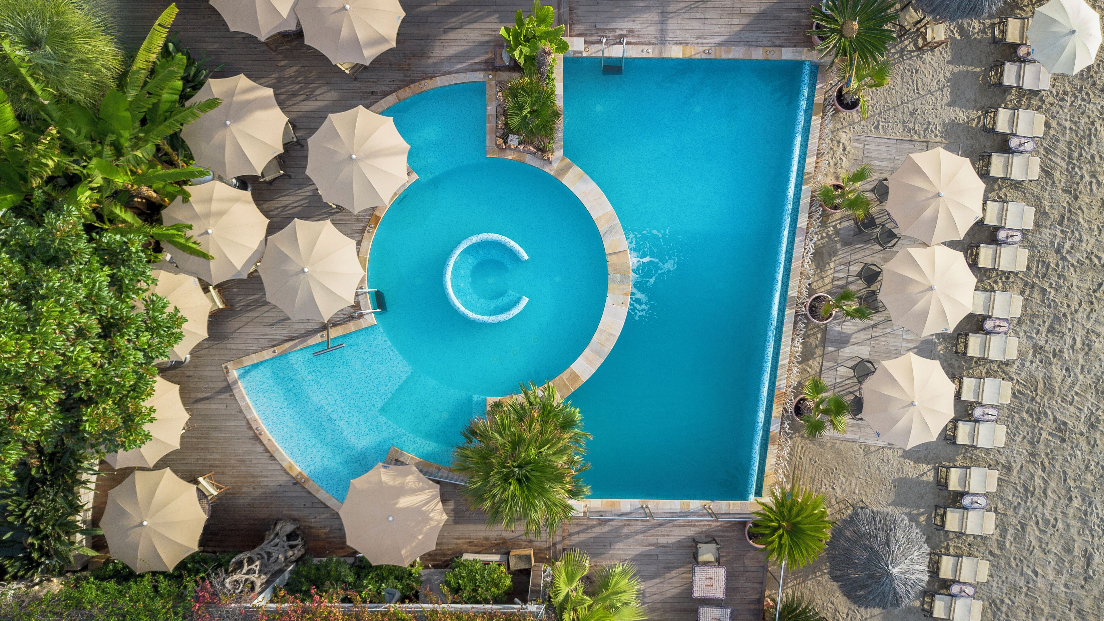 Vista aérea de uma área de piscina com um elemento aquático circular, rodeada por espreguiçadeiras e guarda-sóis.