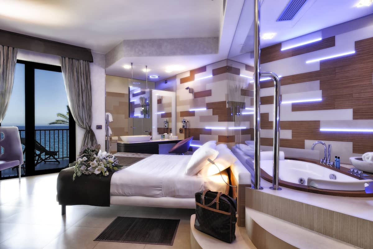A modern hotel room with a large bed, a bathroom with a bathtub, a balcony with a view of the sea, and a desk with a chair.