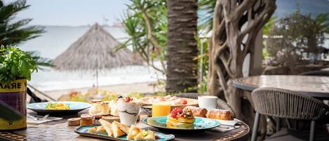 A table set with breakfast items, including pastries, fruits, and beverages, overlooking a beach with palm trees and a thatched hut in the background.