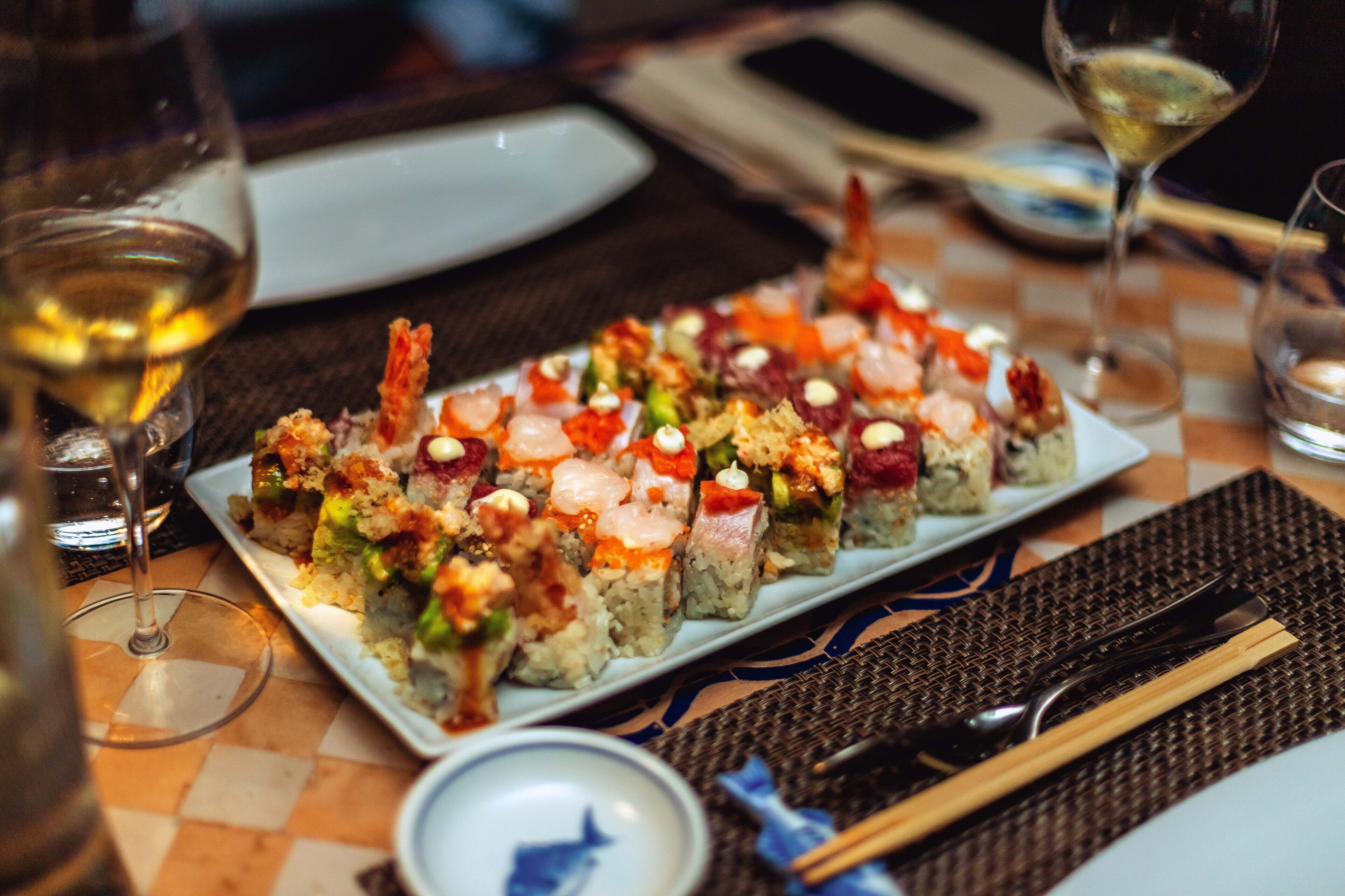 Una bandeja de sushi sobre una mesa con palillos, un tazón pequeño y copas de vino.
