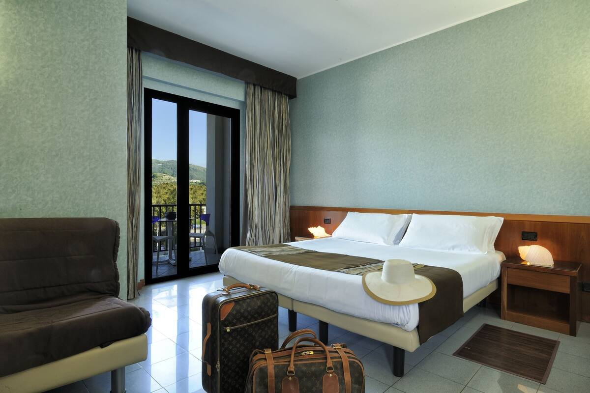 A hotel room with a bed, a sofa, a suitcase, a hat, and a nightstand with a lamp.