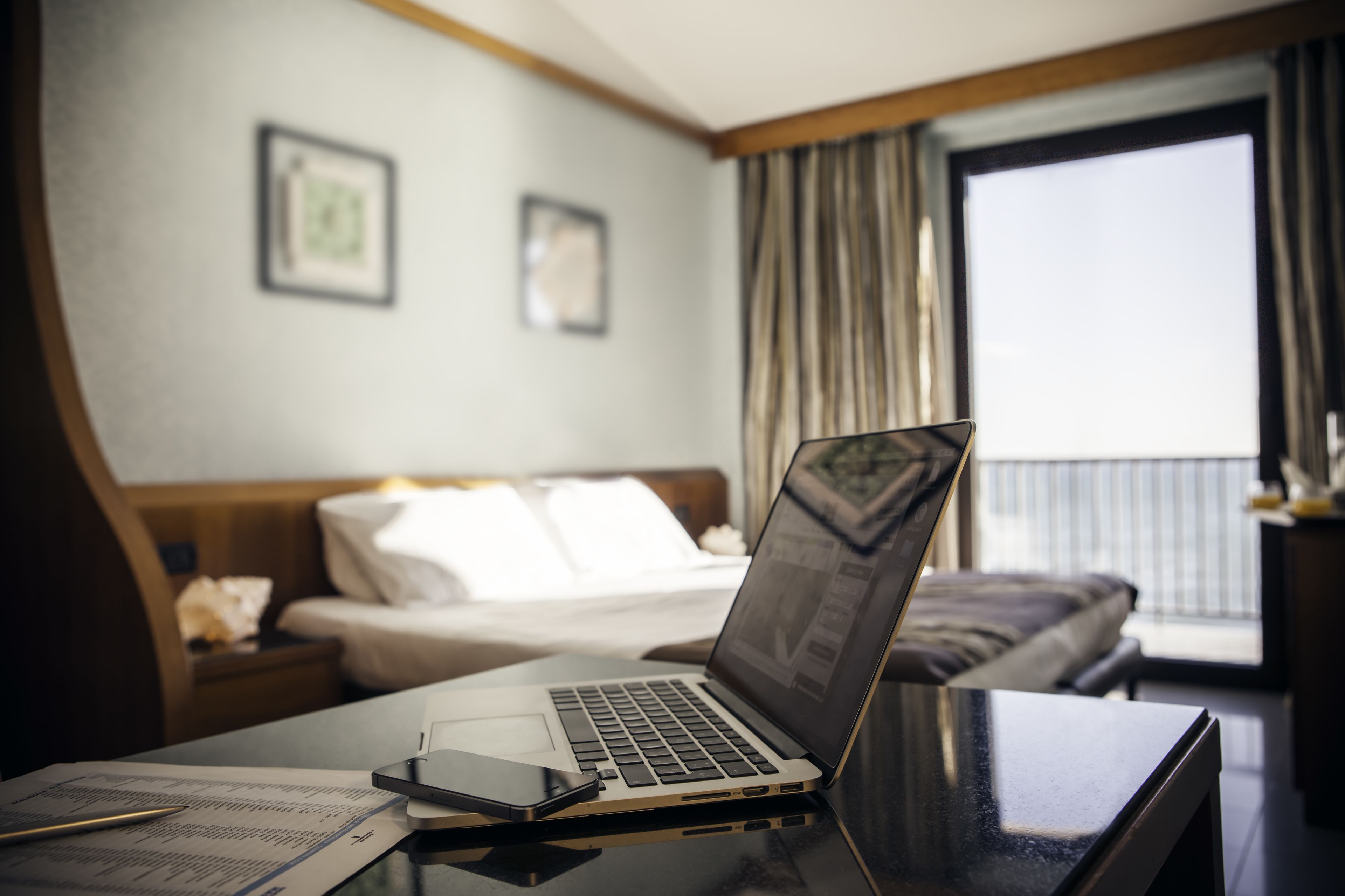 A hotel room with a bed, a desk with a laptop, a bedside table with a lamp, and a window with curtains.