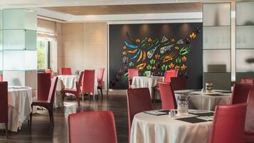 Ein Restaurant mit roten Stühlen und weißen Tischdecken, einer schwarzen Wand mit farbenfrohen Kunstwerken und einer Glasvitrine.