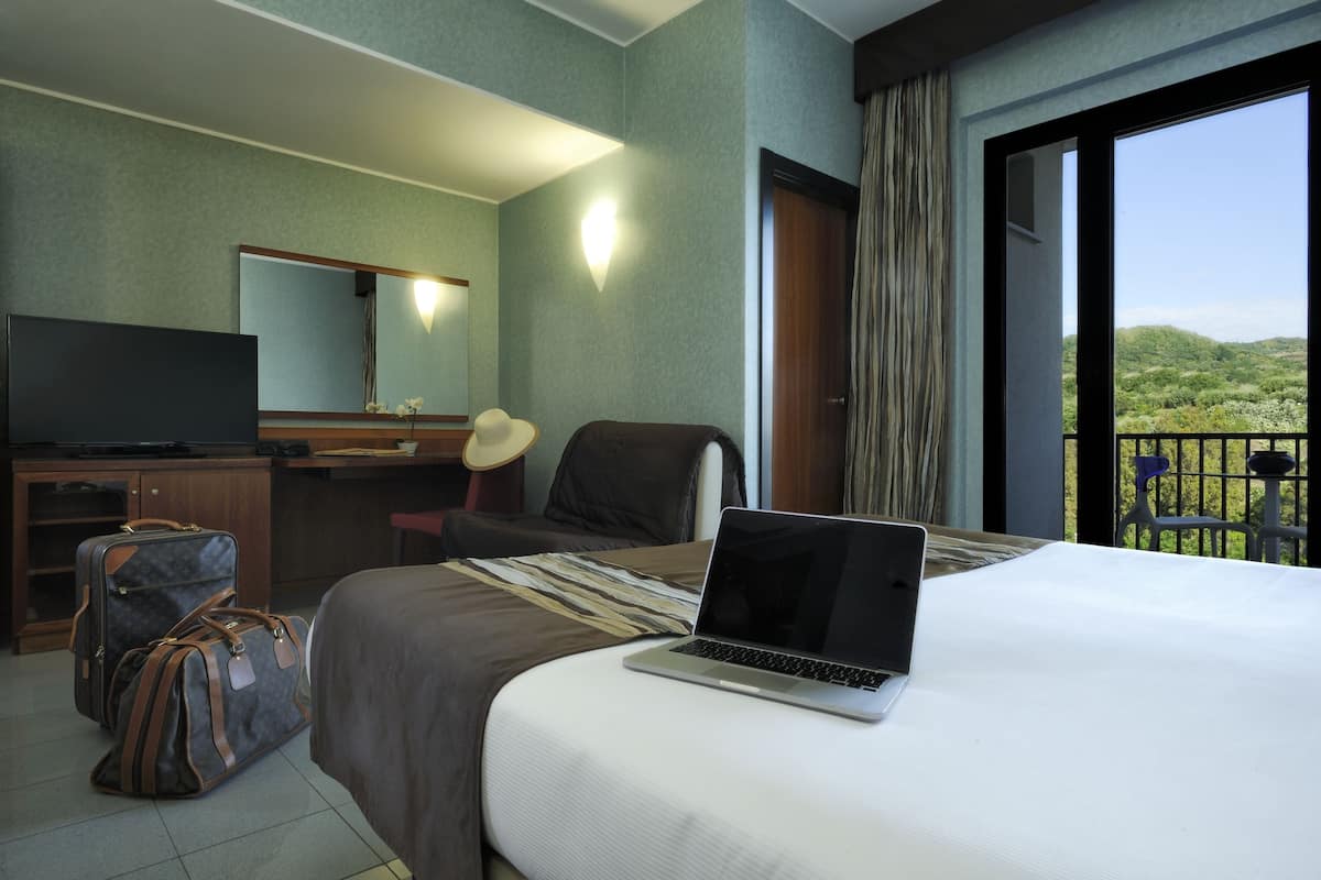 A hotel room with a bed, a desk, a chair, a laptop, and a suitcase.