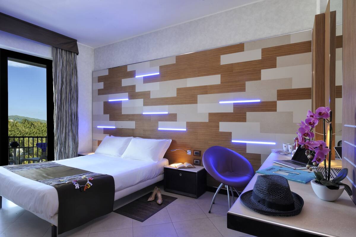 A modern hotel room with a large bed, a desk with a laptop, a chair, and a view of greenery outside.