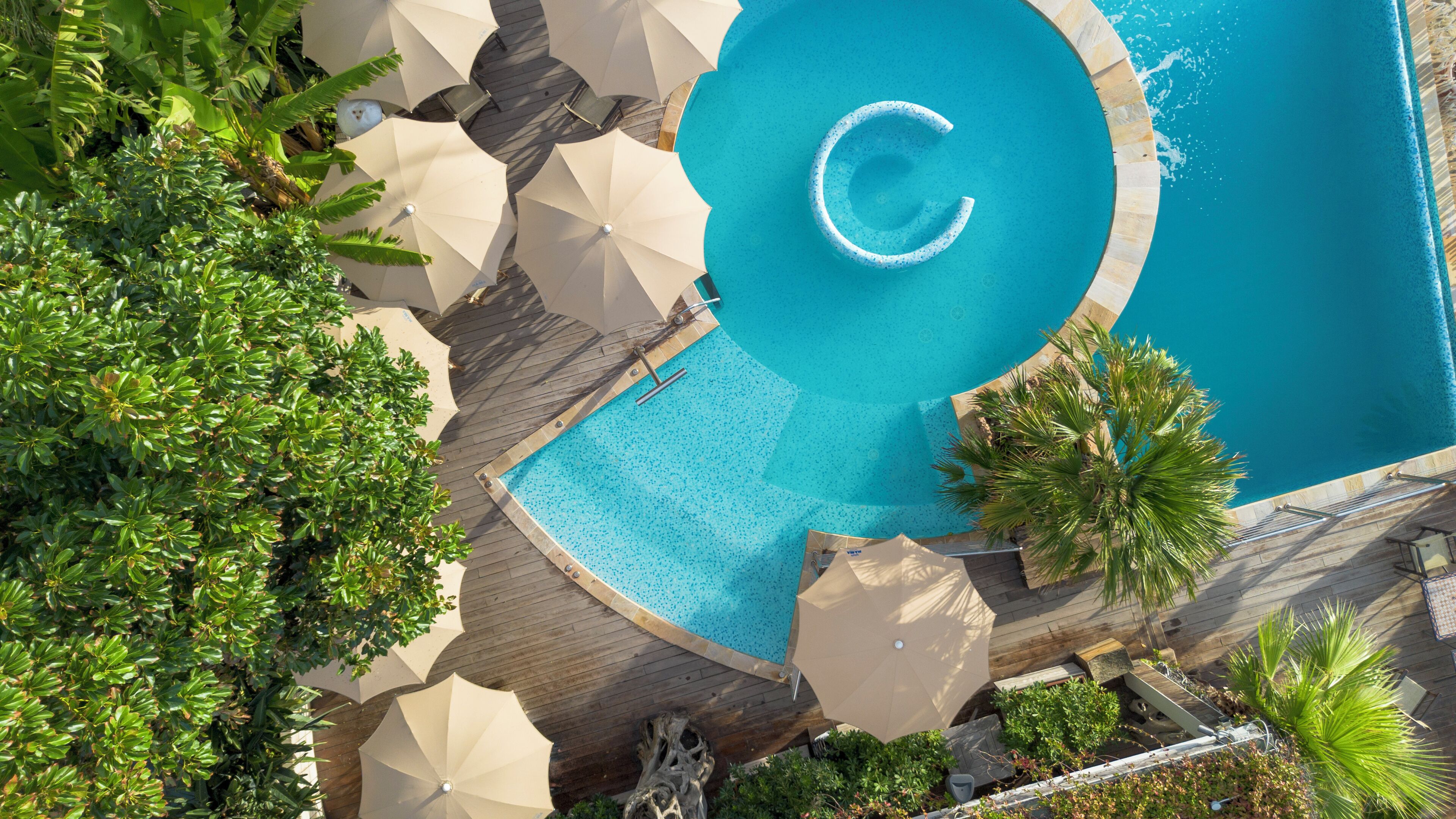 An aerial view of a swimming pool with a circular design, surrounded by palm trees and umbrellas.
