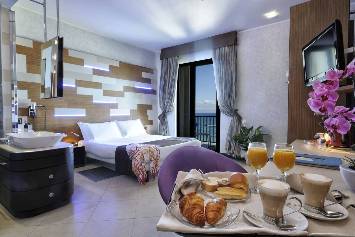 A modern hotel room with a bed, bedside table, a bathroom with a sink, a balcony with a view, and breakfast items on a tray.