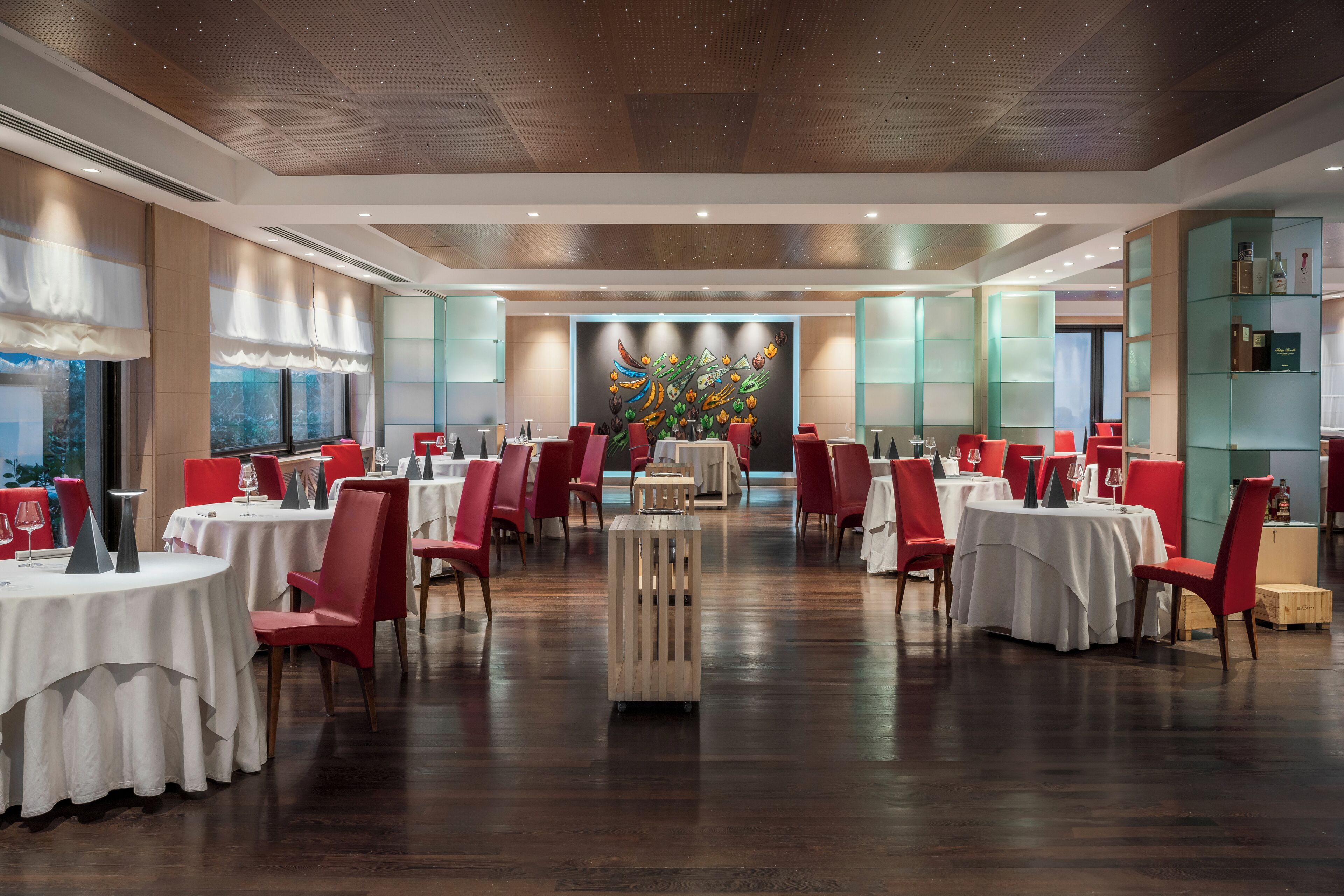 A spacious dining area with round tables and red chairs, a central decorative wall, and a view of the outdoors.