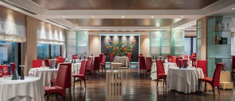 A spacious dining area with round tables and red chairs, a central decorative wall, and a view of the outdoors.