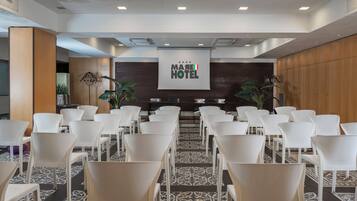 A conference room with rows of white chairs, a large screen displaying "MARE HOTEL" logo, and a patterned carpet.